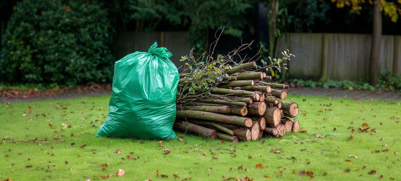 Un sac vert de déchets de jardin est posé à côté d’un tas de branches fraîchement coupées sur une pelouse, illustrant le ramassage et le tri des déchets verts après l’entretien du jardin